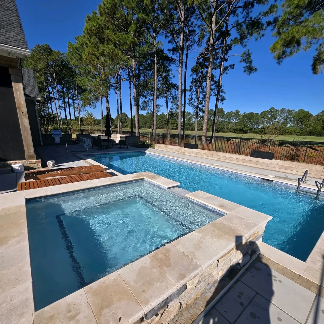 An outdoor swimming pool and hot tub next to a house, surrounded by pine trees with a golf course in the background.
